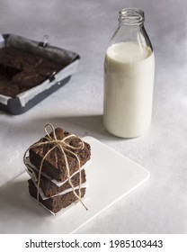 We see a stack of three brownies wrapped and served on a white plate  in the background, we can see a bottle of milk 
