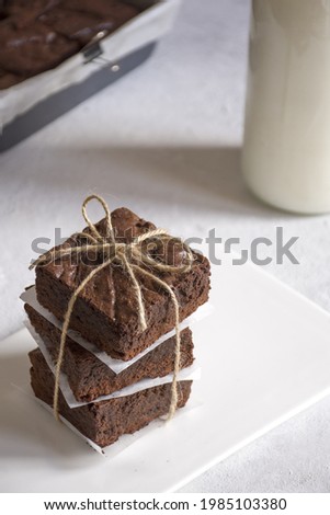 We see a stack of three brownies wrapped and served on a white plate  in the background, we can see a bottle of milk 