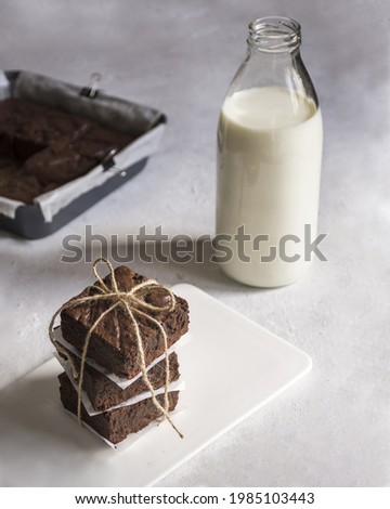 We see a stack of three brownies wrapped and served on a white plate  in the background, we can see a bottle of milk 