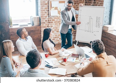 We should use this strategy! Top view of handsome young man in glasses standing near whiteboard and pointing on the chart while his coworkers listening and sitting at the table in office