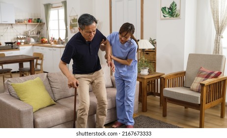 Weak Asian Japanese Older Man Suffering Arthritis Getting Up From Couch Slowly With His Caregiver’s Help In Living Room At Home. His Hand Holding A Stick Is Shaking While Standing