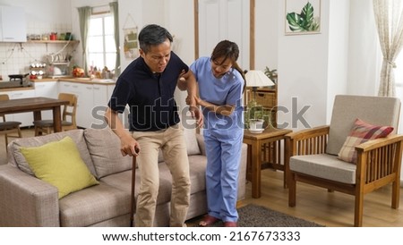 weak asian Japanese older man suffering Arthritis getting up from couch slowly with his caregiver’s help in living room at home. his hand holding a stick is shaking while standing