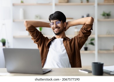 Wealthy Indian Guy Project Manager Sitting At Worktable In Front Of Modern Laptop, Leaning Back And Smiling, Feeling Good About His Job, Office Interior. IT, Programmer, Developer Concept