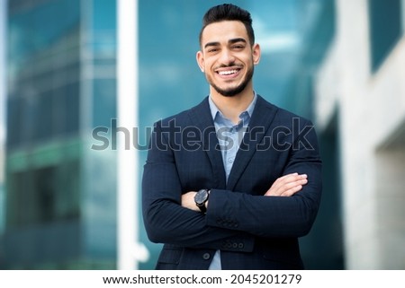 Wealthy middle-eastern young businessman in stylish suit posing next to office building, smiling arab entrepreneur having break after successful busienss meeting, standing on street, copy space