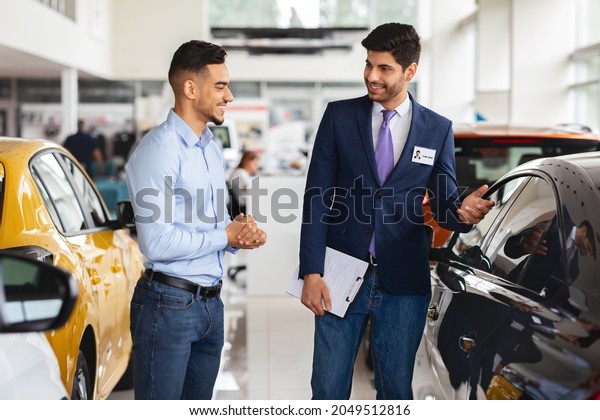 Wealthy middle-eastern\
young man choosing new car, having conversation with friendly sales\
manager at brand new vip showroom, looking at black sports auto and\
smiling, side view