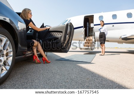 wealthy woman stepping out of car parked in front of private plane and airhostess