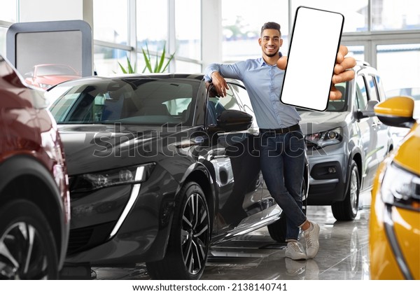 Wealthy young middle eastern man posing by brand\
new black sports car, showing smartphone with empty screen and\
smiling at camera, mockup, renting auto on Internet, copy space.\
Car dealership online