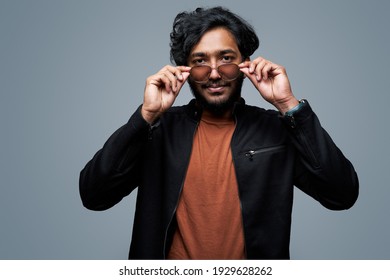 Wearing Black Jacket And Orange Shirt Hipster Person Takes Off His Sunglasses Looking At Camera And Posing In Gray Background.