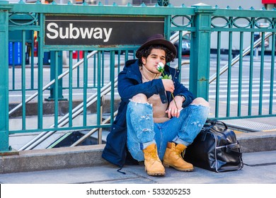 Wearing Jacket With Hood, Destroyed Jeans, Boot Shoes, Fedora Hat, Leather Bag On Ground, Man With Freckle Face, Sits On Street By Subway Sign In New York, Smelling White Rose. Color Filtered Effect