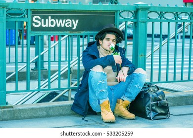 Wearing Jacket With Hood, Destroyed Jeans, Boot Shoes, Fedora Hat, Leather Bag On Ground, Man With Freckle Face, Sits On Street By Subway Sign In New York, Smelling White Rose. Color Filtered Effect