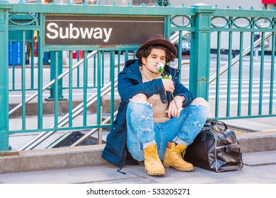 Wearing Jacket With Hood, Destroyed Jeans, Boot Shoes, Fedora Hat, Leather Bag On Ground, Man With Freckle Face, Sits On Street By Subway Sign In New York, Smelling White Rose. Color Filtered Effect