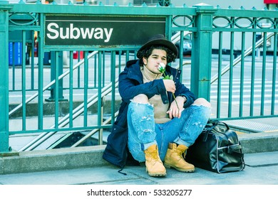 Wearing Jacket With Hood, Destroyed Jeans, Boot Shoes, Fedora Hat, Leather Bag On Ground, Man With Freckle Face, Sits On Street By Subway Sign In New York, Smelling White Rose. Color Filtered Effect