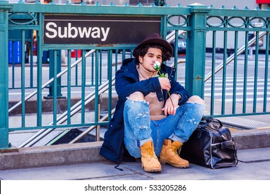 Wearing Jacket With Hood, Destroyed Jeans, Boot Shoes, Fedora Hat, Leather Bag On Ground, Man With Freckle Face, Sits On Street By Subway Sign In New York, Smelling White Rose. Color Filtered Effect