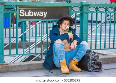 Wearing Jacket With Hood, Destroyed Jeans, Boot Shoes, Fedora Hat, Leather Bag On Ground, Man With Freckle Face, Sits On Street By Subway Sign In New York, Smelling White Rose.