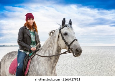 Wearing Jeans, Jacket And Fall Hat Female Rider Goes On Horseback Along Sandy Seabeach