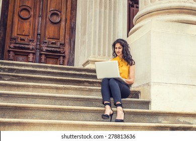 Wearing Sleeveless Orange Shirt, Striped Pants, High Heels, A Young East Indian American College Student Sitting On Stairs Outside Office Building On Campus, Working On Laptop Computer, Reading.
