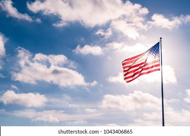 Weather Clouds And Sunshine Summer Sky With American Flag Illuminated Back Lit By Sun. United States Flag Waving In The Wind On A Dramatic Sky Day