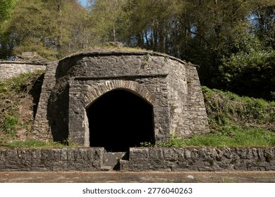 A weathered, ancient stone structure, possibly a lime kiln or ice house, set into a grassy hillside with trees overhead, showcasing signs of age and exposure to the elements.