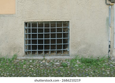 A weathered barred window set into a rough textured wall with overgrown weeds and stones at its base under natural daylight offering a glimpse of an unseen interior space