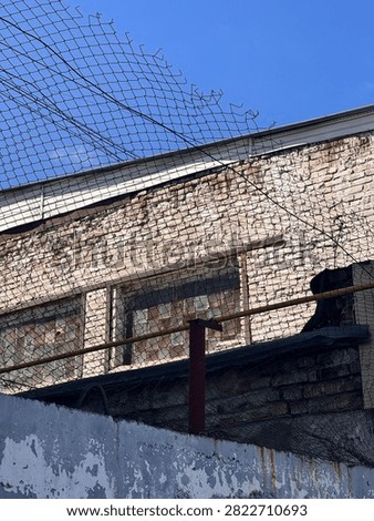 Weathered brick building viewed through damaged chain-link fence against clear blue sky, showing urban decay and deterioration.