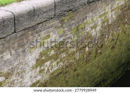 Weathered Brick Canal Wall Dated 1909 Covered In Moss And Algae With Waterline Stains