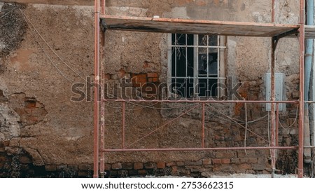 Weathered brick and plaster wall of an old building with a broken window and metal scaffolding during renovation work. Detailed view of urban decay and restoration process in historic architecture.