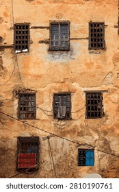 A weathered building facade with multiple windows, some with decorative ironwork, shows signs of age and disrepair, with peeling paint and exposed brick, while various objects like a towel and antennas are placed in or near the windows.