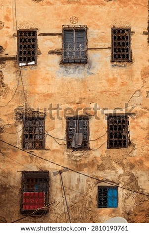 A weathered building facade with multiple windows, some with decorative ironwork, shows signs of age and disrepair, with peeling paint and exposed brick, while various objects like a towel and antennas are placed in or near the windows.