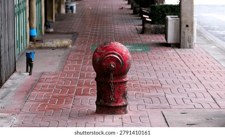 A weathered cast iron fire hydrant with peeling red paint, rust patches, and a small hanging chain stands on a wide decorative brick-patterned sidewalk in Bangkok, with a long pedestrian path receding into a soft bokeh background of benches and greenery.