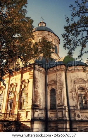 A weathered church with peeling paint stands amidst lush trees under a clear sky. The Church of St. John the Evangelist is an Orthodox church and an architectural monument in Nizhyn.