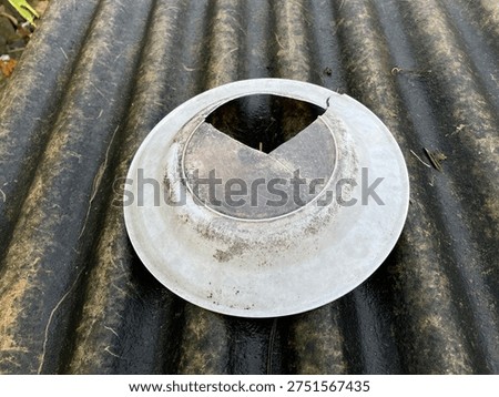 A weathered, circular white plastic light fixture cover or utility cap with a broken triangular section, resting on a dark, mossy corrugated roofing sheet. The image highlights themes of decay, abandoned construction, and weathered outdoor materials.