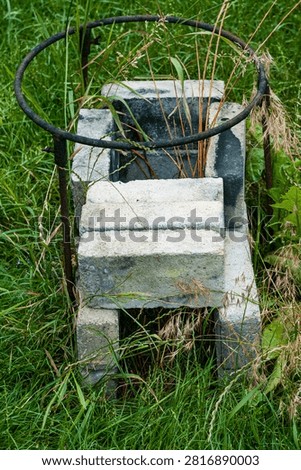 A weathered concrete block structure with a rusted metal ring enclosure is surrounded by tall green grass and wild plants in an outdoor setting.