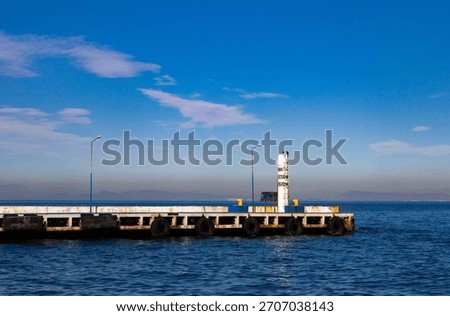 Weathered Concrete Jetty Structure Lined with Old Rubber Tire Fenders and Featuring a Small White Navigation Beacon Light, Extending into Calm Blue Sea Under a Sunny Sky with Distant Horizons.