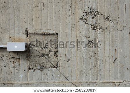 Weathered concrete wall features a rectangular patch with exposed rebar and an electrical box with a visible wire. The rough surface adds an industrial feel to the scene.