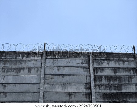 A weathered concrete wall with multiple strands of barbed wire running along the top. The wall is set against a clear blue sky.