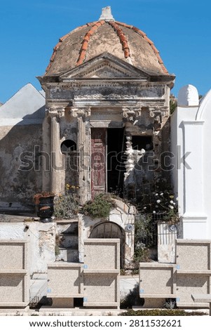 A weathered domed mausoleum with classical columns and a crumbling facade stands amid a Mediterranean cemetery.