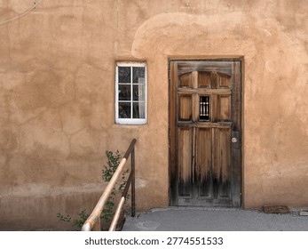 A weathered door and tiny window are set into an ancient adobe facade. Chimayo NM 2026.