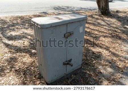 Weathered gray electrical utility box, or telecommunications enclosure, standing securely beside a paved asphalt road, surrounded by dry oak leaves and dappled tree shadows on a sunny day, symbolizing essential public infrastructure.