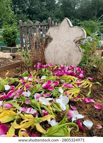 A weathered, light-colored tombstone with a distinctive, rounded shape stands amidst a bed of rich, dark earth, adorned with a vibrant scattering of pink, white, 
