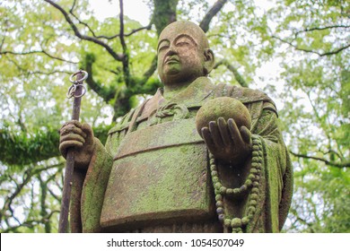 A weathered, moss covered, life size stone statue of a Buddhist monk holding a walking staff and wearing Buddhist prayer beads meditates under green tree leaves in a forest in Japan.