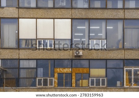 Weathered office facade with windows and panels, worn concrete exterior shows air vents, goldtoned entrance and grid of glass reflecting muted sky, utilitarian