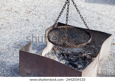A weathered outdoor fire pit with a hanging grill grate, poised over ash and char, captures the essence of rustic barbecue and campfire cooking.