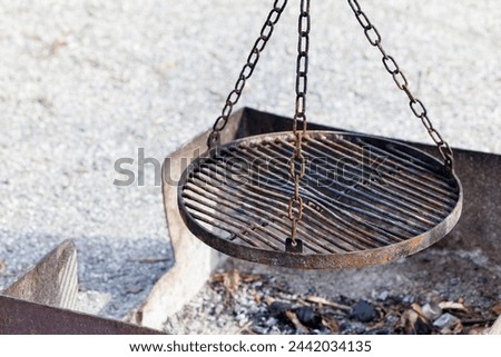 A weathered outdoor fire pit with a hanging grill grate, poised over ash and char, captures the essence of rustic barbecue and campfire cooking.