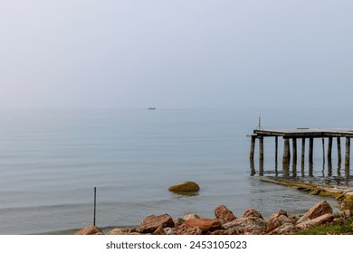 A weathered pier juts into the placid waters of Lake Garda, while a solitary boat on the horizon drifts through the fog on a quiet Italian morning - Powered by Shutterstock