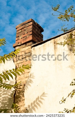 A weathered red brick chimney stands atop a worn, cracked white wall. Sunlight casts shadows from nearby tree leaves, against a clear blue sky. Rustic and timeless.