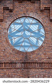 Weathered red brick facade with large circular window, geometric glass panes, and arched masonry framing, showing symmetry, texture, and historic industrial architecture detail in soft daylight