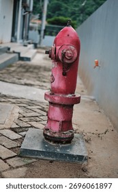 A weathered red fire hydrant standing on a concrete block next to a gray wall. The bright, vivid color contrasts with the subdued urban environment, suggesting themes of emergency, safety, and infrast