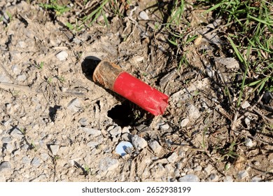 A weathered red shotgun shell casing lies abandoned in the dirt and gravel, partially buried and rusted, highlighting environmental pollution and human impact on natural areas