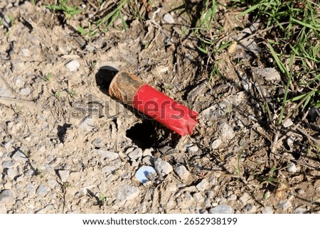 A weathered red shotgun shell casing lies abandoned in the dirt and gravel, partially buried and rusted, highlighting environmental pollution and human impact on natural areas
