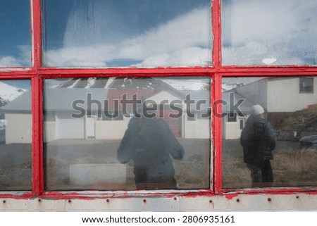 A weathered red-framed window in Dalvik, North Iceland, reflects harbor houses and snow-capped mountains alongside the silhouette of a photographer in a quiet spring moment.
